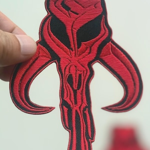 Pode incluir: Patch bordado com um design de caveira vermelha e preta com chifres curvos. O patch tem uma aparência detalhada e texturizada, com o design delineado em preto. O patch é segurado por uma mão contra um fundo branco.