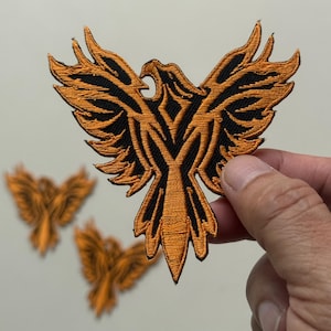 Può includere: Toppa ricamata con un design stilizzato di fenice con le ali spiegate. La toppa ha una combinazione di colori nero e arancione, con il corpo e le ali della fenice dettagliati con cuciture intricate. Sono visibili toppe aggiuntive sullo sfondo.