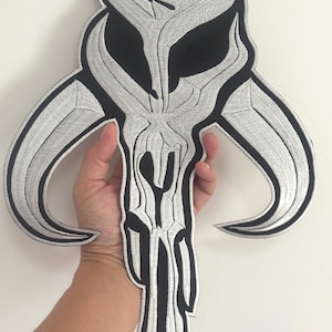 Peut inclure: Écusson brodé avec un motif de crâne stylisé en noir et argent. Le patch a un aspect texturé et détaillé avec une forme symétrique. Le design comprend des cornes incurvées et un élément vertical central.