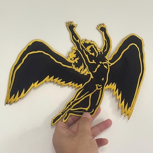 Puede incluir: Parche bordado en negro y dorado que representa una figura alada con los brazos levantados. El cabello y el contorno de la figura son dorados, con alas negras bordeadas en dorado. El parche se sostiene sobre un fondo blanco.