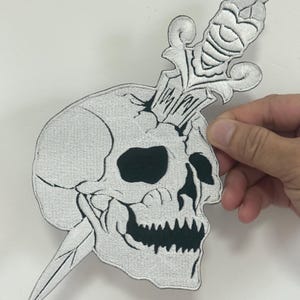 May include: Embroidered patch featuring a skull pierced by a decorative dagger. The skull is white with black eye sockets and a jagged mouth. The dagger has an ornate handle and a sharp blade. The patch is likely for clothing or accessories.