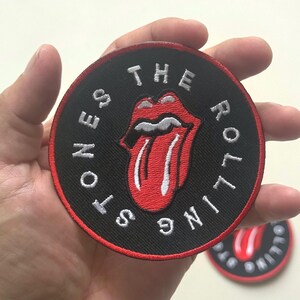 May include: A round black patch with the words "THE ROLLING STONES" in white lettering around the edge. The patch features the iconic red tongue and lips logo. A second patch is visible in the background.