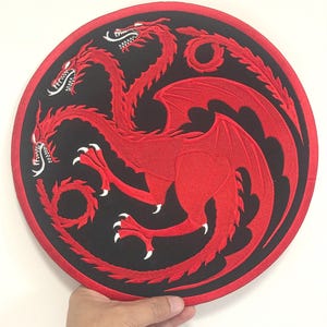 Puede incluir: Parche bordado con un diseño de dragón rojo de tres cabezas sobre un fondo circular negro. El dragón tiene alas detalladas y garras afiladas. El parche tiene un borde rojo.