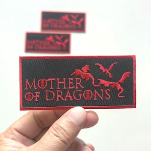 以下が含まれることがあります： 赤い刺繍のテキストとドラゴンのシルエットが入った黒い長方形のパッチ。「MOTHER OF DRAGONS 2」と書かれています。パッチには赤い縁取りがあり、手で持っています。背景にはさらに2つのパッチが見えます。