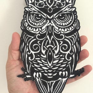 May include: A black and white embroidered owl patch with detailed patterns. The owl design includes feathers and facial features. The patch is held in a hand.