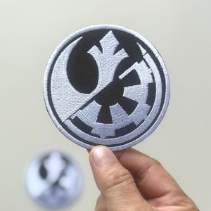 Peut inclure: Écusson brodé rond avec un design divisé. Une moitié présente un symbole blanc de l'Alliance Rebelle, l'autre un symbole impérial noir, sur fond noir. L'écusson a une bordure blanche. Un second écusson flou est en arrière-plan.