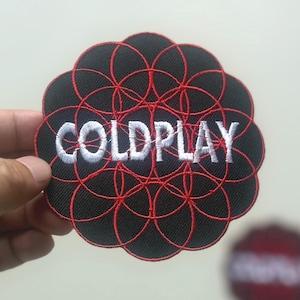 May include: Black patch with the word "COLDPLAY" in white, embroidered over a red circular design. The patch has a scalloped edge. A second, blurred patch is visible in the background.