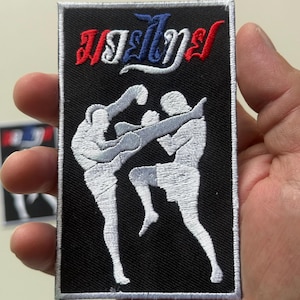 Peut inclure: Écusson rectangulaire noir avec un motif Muay Thai. L'écusson présente du texte thaï en rouge, blanc et bleu. Deux figures blanches sont représentées en action, engagées dans un combat de Muay Thai. L'écusson est tenu par une main.