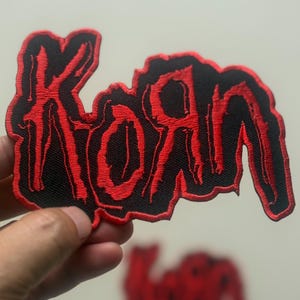 May include: A black patch with the word "KORN" embroidered in red, stylized with a dripping effect. The patch is held up against a light background, with a second, smaller patch visible in the background.
