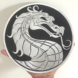 Peut inclure: Un écusson rond noir avec un motif de dragon blanc brodé. Le dragon est représenté dans une posture incurvée, avec sa tête et sa queue se touchant. L'écusson est tenu par une main.