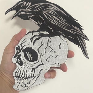 Può includere: Toppa ricamata raffigurante un corvo nero appollaiato su un teschio. Il corvo e il teschio sono dettagliati con cuciture bianche. Il teschio ha un aspetto strutturato bianco sporco. La toppa è progettata per l'applicazione su abbigliamento o accessori.