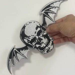 May include: Embroidered patch depicting a skull with bat wings. The skull has detailed shading, and the wings are black with silver edging. The patch is held against a white backdrop.