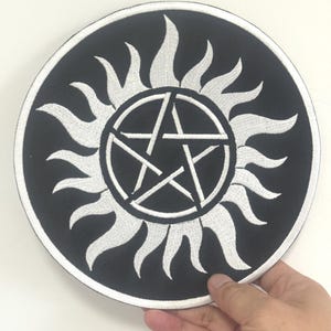 Peut inclure: Écusson rond noir avec un motif de soleil blanc entourant un pentagramme blanc. Le patch est tenu par une main sur un fond blanc. Le motif est brodé.