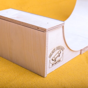 Pode incluir: Rampa de skate de madeira clara com uma superfície curva e uma base retangular. A rampa tem as palavras "SKATE DREAMS" e um gráfico de skate gravado na lateral. A rampa está sobre uma superfície amarela.