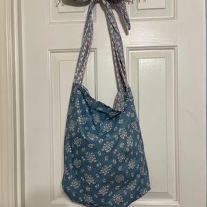 May include: A blue tote bag with a white floral pattern hangs from a white metal rack on a white door. The bag's straps and the bow holding the rack are patterned with red and blue flowers on a white background.