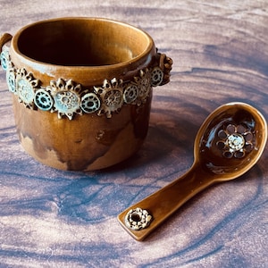 Könnte beinhalten: Ein braunes Keramikschalen- und Löffel-Set. Die Schale hat ein dekoratives Band aus blauen und braunen floralen Designs um den Rand. Der Löffel hat ein ähnliches florales Design in der Schale und am Griff.