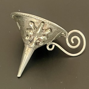 May include: A small, silver-colored funnel with a decorative floral design on the bowl. The funnel has a long, tapered spout and a curled handle. The object is likely used for pouring liquids or powders.