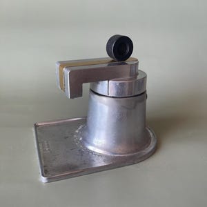 Può includere: Una caffettiera in metallo color argento con base rettangolare piatta, corpo cilindrico e braccio orizzontale con pomello circolare nero sulla parte superiore. La base presenta la scritta in rilievo "2 TAZZE" e "BREVETTATO".