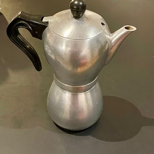 Può includere: Una caffettiera espresso argentata con manico e pomello del coperchio neri. La caffettiera ha una base rotonda e una camera superiore bulbosa con un beccuccio. Il design è in stile classico e vintage.