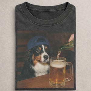 May include: A charcoal gray t-shirt featuring a graphic print of a tricolor dog wearing a blue baseball cap, sitting at a table while a liquid is poured from a glass bottle into a clear glass mug filled with a golden amber beverage.