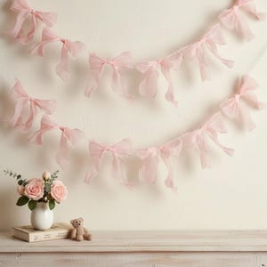 May include: Two strands of sheer pink ribbon bows are strung across a white wall. A small vase of pink roses, a book, and a teddy bear sit on a wooden surface below the bows. The bows are delicate and airy.