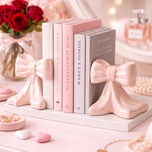 May include: A set of pink ceramic bow-shaped bookends holding a row of books in shades of pink and gray. The books are on a white shelf, with a pink surface below. Decorative items like rings, hearts, and a vase of roses complete the scene.