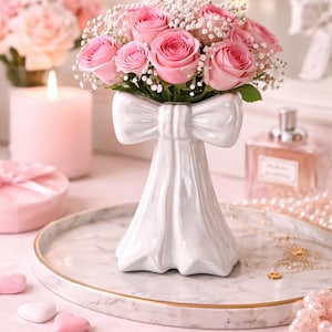 Può includere: Un vaso in ceramica bianca a forma di abito con fiocco, contenente un bouquet di rose rosa e velo da sposa. Il vaso è su un vassoio di marmo con bordi dorati, circondato da decorazioni rosa e perle.