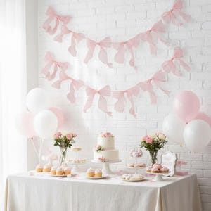 May include: A baby shower table setting with a white tablecloth, a two-tiered cake, cupcakes, and pink and white balloons. Pink bow garlands hang on the white brick wall. The table is decorated with flowers and treats.