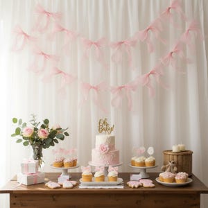 May include: A baby shower dessert table with a two-tiered pink cake, cupcakes, and cookies. A garland of pink bows hangs above the table. The cake is topped with a gold "Oh Baby!" cake topper. A vase of pink roses and a gift box are also on the table.