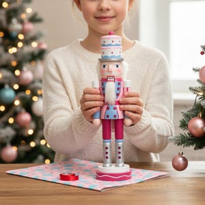 May include: A pink, blue, and white wooden nutcracker doll with a tall hat and a long white beard. The nutcracker is held by a person in front of a Christmas tree with pink and blue ornaments. The doll is approximately 12 inches tall.