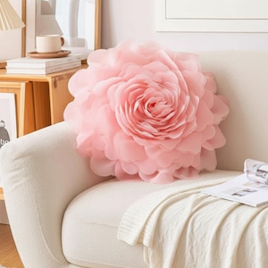 Könnte beinhalten: Ein großes, hellrosa Zierkissen in Rosenform mit mehreren Blütenblätterschichten. Das Kissen liegt auf einem cremefarbenen Sofa und schafft eine weiche und einladende Ästhetik. Das Kissen verleiht dem Wohnraum einen Hauch floraler Eleganz.
