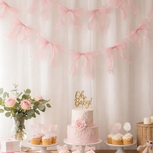 May include: A beautifully decorated table for a baby shower, featuring a two-tiered cake with a gold "Oh Baby!" topper, cupcakes, cookies, and pink bow garlands. A vase of pink roses and gift boxes add to the festive decor.