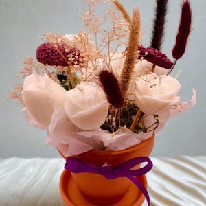 May include: A small terracotta pot filled with white, cream, and burgundy-colored flowers and decorative stems. The pot is tied with a purple ribbon. The arrangement includes white rose-shaped candles.