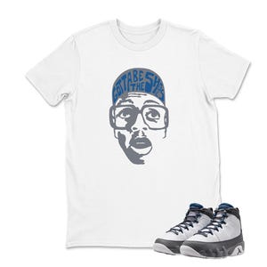 May include: White t-shirt featuring a graphic of a person wearing glasses with the text "Gotta Be The Shoes" on a blue hat. A pair of white, black, and blue sneakers are in the foreground.