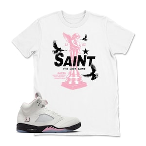 May include: White t-shirt with a pink and black graphic of a saint and birds, paired with white and black sneakers with pink accents. The shirt features the text "SAINT THE LOST SAINT". The sneakers have the number "23" on the side.