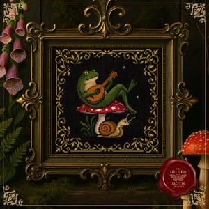 Puede incluir: Arte de punto de cruz en un marco dorado. La obra representa una rana verde tocando un laúd sobre una seta roja y blanca, con un caracol debajo. El fondo es negro con detalles florales dorados. El marco tiene un acabado verde oscuro.