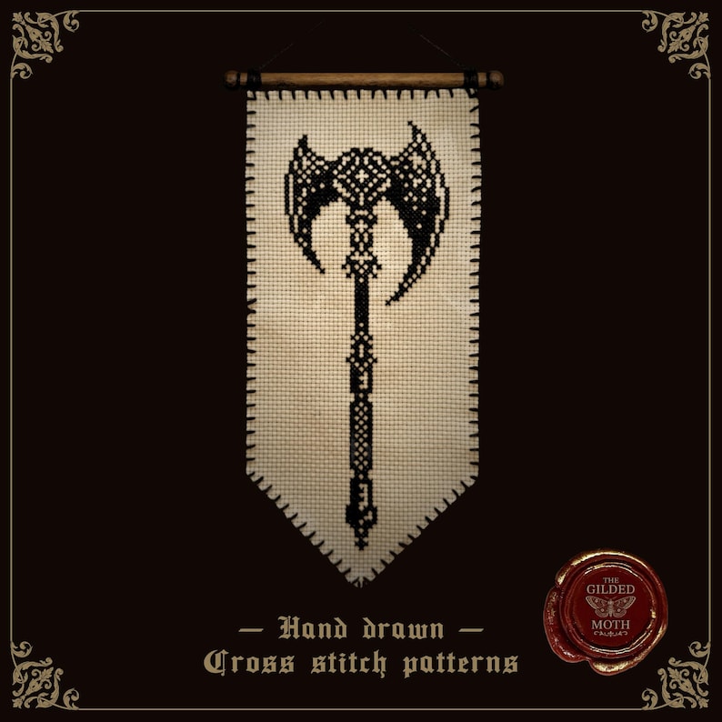 May include: A cream-colored fabric banner featuring a black cross-stitch design of a double-headed battle axe. The banner is suspended from a wooden rod and includes the text "Hand drawn Cross stitch patterns" and a red wax seal with "THE GILDED MOTH".
