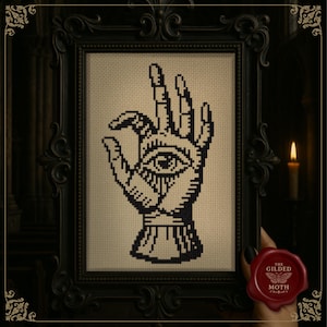 以下が含まれることがあります： 黒いフレームに入ったクロスステッチアート。手のひらに目がある手を描いています。アートワークは、装飾的なディテールのある黒いフレームに入っています。背景はニュートラルカラーです。「The Gilded Moth」と書かれた赤いワックスシールが見えます。