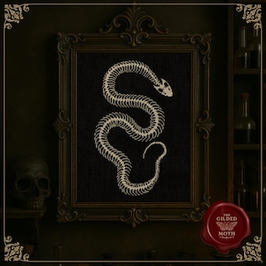 May include: A framed artwork featuring a snake skeleton design in off-white against a black background. The frame is ornate and dark, with a skull visible on the left. The artwork is from "The Gilded Moth".