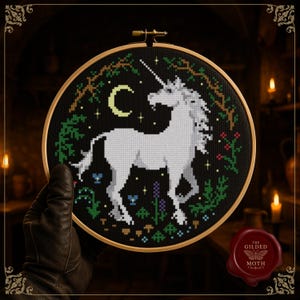 May include: A cross-stitch embroidery hoop showing a white unicorn on a black backdrop with a crescent moon and stars. The design includes green foliage and flowers. The hoop is held by a gloved hand. The Gilded Moth logo is in the bottom right.