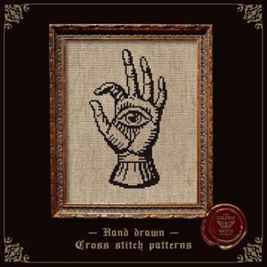 May include: A cross stitch pattern featuring a black graphic of a hand with an eye in the palm, displayed in a rectangular ornate gold frame. Below the frame, the text reads: Hand drawn Cross stitch patterns, with a wax seal logo for The Gilded Moth.