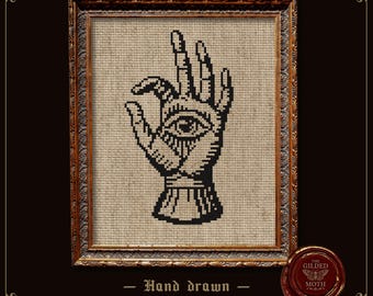 May include: A cross stitch pattern featuring a black graphic of a hand with an eye in the palm, displayed in a rectangular ornate gold frame. Below the frame, the text reads: Hand drawn Cross stitch patterns, with a wax seal logo for The Gilded Moth.