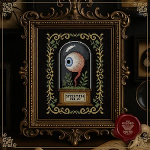 Könnte beinhalten: Kreuzstich-Kunstwerk eines Augapfels in einer Glaskuppel, beschriftet mit "Specimen No. IV". Das gerahmte Stück hat einen schwarzen Hintergrund mit grünen und goldenen floralen Akzenten. Der Rahmen ist verziert, goldfarben und im Vintage-Stil.