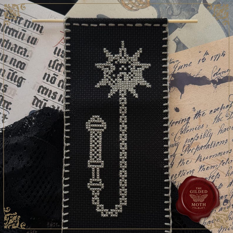 May include: Black cross-stitch bookmark featuring a stylized sunburst design and a decorative handle. The bookmark is stitched with white thread on black fabric, with a white stitched border. The background includes vintage paper and a wax seal.