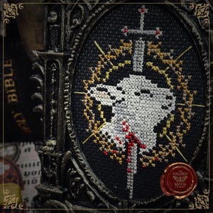 May include: A framed cross-stitch design featuring a white skull pierced by a silver sword with a red cross. The skull is surrounded by a gold and yellow wreath on a black background. The frame is ornate and dark.