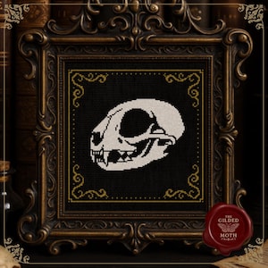 May include: A framed cross-stitch depicting a white cat skull against a black backdrop. The frame is elaborately designed, with a dark bronze finish and gold detailing. A red wax seal with "THE GILDED MOTH" is visible. The style is gothic and vintage, with a macabre theme.