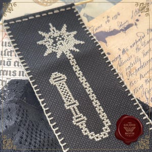 May include: Black and white cross-stitched bookmark featuring a mace design. The bookmark has a white stitched border and is set against a backdrop of aged paper and lace. A red wax seal with a butterfly is in the lower right corner.