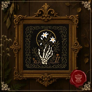 May include: Cross-stitch art in a gold ornate frame. The artwork features a skeletal hand holding white flowers against a black background. A yellow halo surrounds the flowers. The frame has a red wax seal with the text "The Gilded Moth".