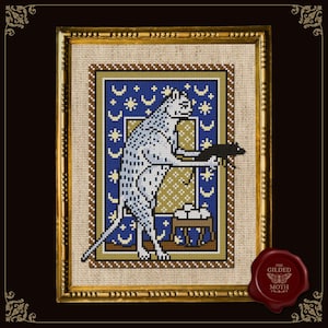 Op de afbeelding: Kruissteekkunstwerk van een witte kat met zwarte vlekken die een zwarte muis vasthoudt, tegen een blauwe achtergrond met sterren en manen. Het ingelijste stuk heeft een gouden rand en is gelabeld "The Gilded Moth".
