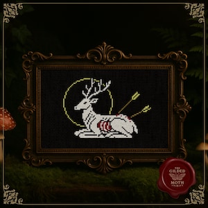 Può includere: Un'opera d'arte a punto croce incorniciata raffigurante un cervo bianco con frecce, su sfondo nero. Il cervo è all'interno di un cerchio giallo. La cornice ornata, marrone scuro, circonda l'opera, esposta su una superficie muschiosa.